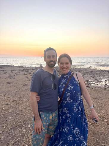 Um casal sorridente está de braços dados em uma praia rochosa ao pôr do sol, com o mar calmo e o céu em tons pastéis ao fundo, irradiando felicidade e calor.