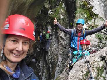 A smiling woman in a red helmet takes a selfie during a rock climbing adventure with family and friends in a scenic, mossy canyon, everyone looks happy.