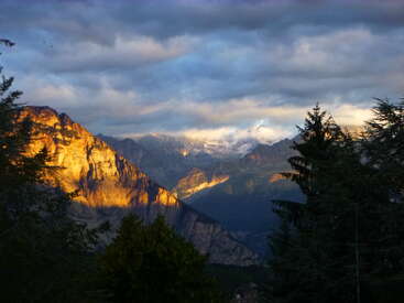 Golden sunlight bathes rugged mountain peaks, partially covered by dramatic clouds. Dark evergreen trees frame the foreground, highlighting the contrast between shadow and glowing rocky cliffs.