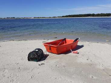 Das Bild zeigt eine ruhige Strandszene mit einem kleinen orangefarbenen Boot und einer schwarzen Krabbenfalle auf dem Sand mit Blick auf ein ruhiges Gewässer.