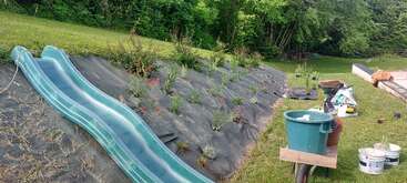 A green plastic slide descends a grassy hill bordered with landscaping fabric, new plants, and gardening supplies. A wheelbarrow, buckets, and garden tools sit nearby.