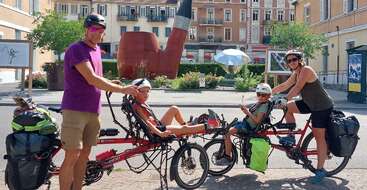 A family of four poses on two tandem recumbent bikes, fully loaded with travel bags. They wear helmets and sunglasses, with a giant pipe sculpture behind them.