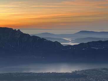A stunning sunrise paints the sky orange above layered mountain ranges, misty valleys, and a tranquil lake, while a small town stretches along the waterfront below.