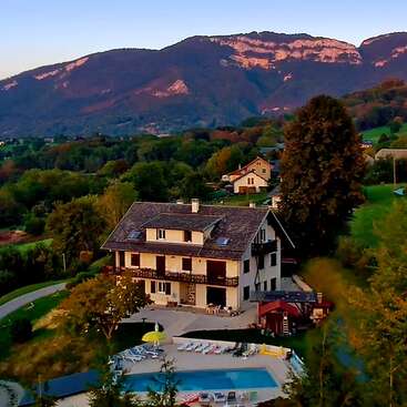 Ein charmantes Landhaus mit Satteldach liegt, umgeben von üppigen Bäumen, einem Swimmingpool und mit Blick auf die Berge, unter einem farbenprächtigen Abendhimmel.
