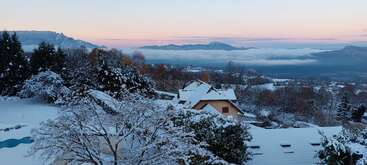 Eine malerische Winterlandschaft zeigt schneebedeckte Häuser und Bäume. Ferne Berge, ein rosafarbener Morgenhimmel und eine Nebelschicht schaffen eine friedliche, ruhige Morgenszene.