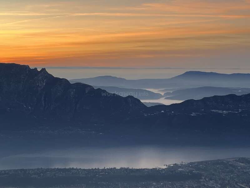 A stunning sunrise paints the sky orange above layered mountain ranges, misty valleys, and a tranquil lake, while a small town stretches along the waterfront below.