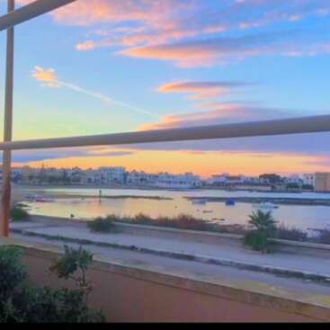 Das Bild zeigt eine ruhige Szene eines Gewässers, mit einer Stadtlandschaft im Hintergrund und einem leuchtenden Himmel bei Sonnenuntergang, von einem Balkon aus betrachtet.
