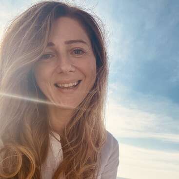 Das Bild zeigt eine Frau mit langen, gewellten Haaren, die in die Kamera lächelt, vor einem blauen Himmel mit weißen Wolken und einer hellen Sonne, die hinter ihr scheint.