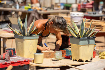 Una mujer trabaja concentrada en un taller al aire libre, rodeada de herramientas, madera y dos grandes plantas de agave en macetas, en un día soleado.
