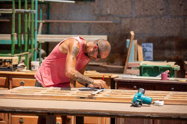 Un hombre vestido con una camiseta sin mangas rosa y gafas de sol está marcando madera en un taller. Herramientas, muebles y materiales le rodean mientras trabaja concentrado al aire libre.