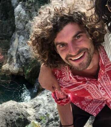 A smiling man with curly hair wearing a red patterned shirt poses outdoors by rocky cliffs and clear water, enjoying nature with a friendly, happy demeanor.