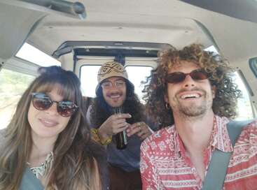 Three friends are enjoying a road trip in a van, smiling and wearing sunglasses. One holds a drink. The atmosphere is relaxed, cheerful, and adventurous.