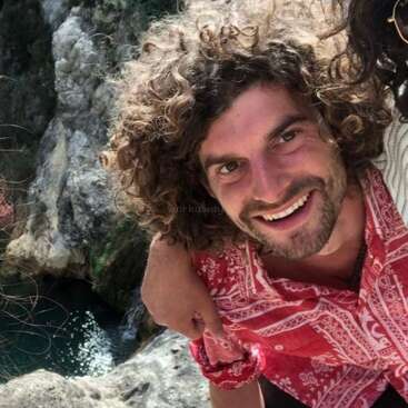 Un homme souriant aux cheveux bouclés, portant une chemise à motifs rouges, pose en plein air près de falaises rocheuses et d'une eau limpide, profitant de la nature avec un comportement amical et heureux.
