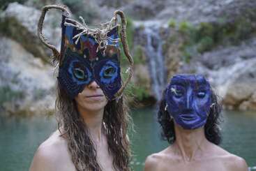 Zwei Leute stehen im Wasser vor einem Wasserfall und tragen jeweils einzigartige, bunte, handgemachte Masken. Die Umgebung ist natürlich, ruhig und ein bisschen geheimnisvoll, was Neugier weckt.