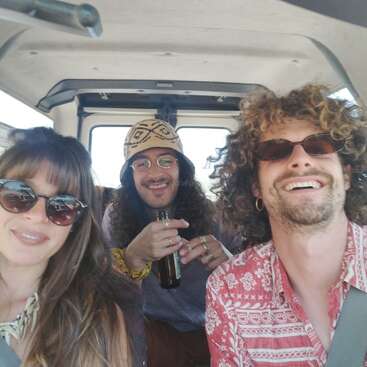 Drei Freunde genießen einen Road Trip in einem Van, lächeln und tragen Sonnenbrillen. Einer hält ein Getränk in der Hand. Die Atmosphäre ist entspannt, fröhlich und abenteuerlich.