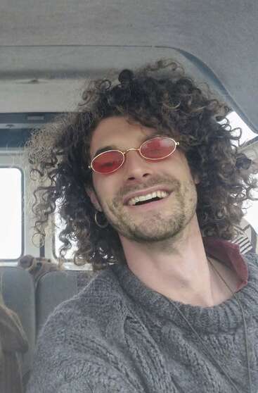 A smiling man with curly hair wears red-tinted sunglasses, a grey knit sweater, and an earring, sitting inside a vehicle, radiating a cheerful, carefree vibe.