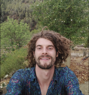 A man with curly hair and a beard is smiling, wearing a colorful patterned shirt, outdoors in a lush, green, wooded area with trees behind him.