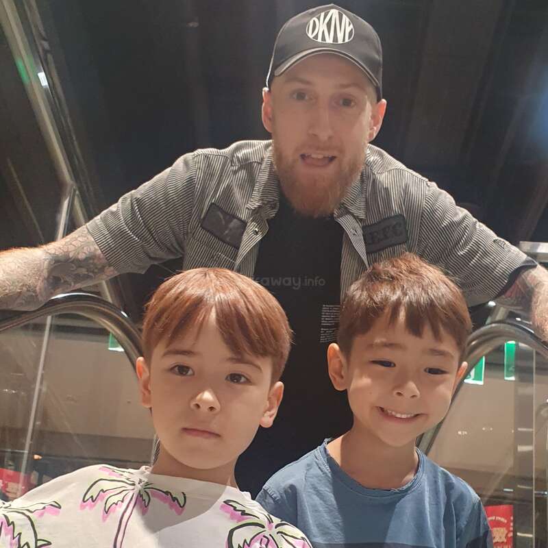 L'image montre un homme posant avec deux jeunes garçons sur un escalator, l'homme portant une casquette et les garçons des vêtements décontractés.