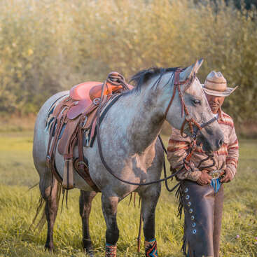 Un vaquero junto a un caballo gris ensillado en un campo cubierto de hierba, con sombrero, zahones y camisa estampada. La escena es tranquila y rústica.