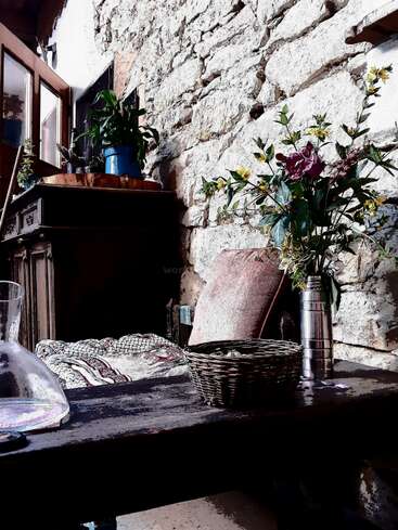 Eine gemütliche, rustikale Ecke mit einer Steinwand, Holzmöbeln, Topfpflanzen, bunten Kissen, einem geflochtenen Korb, einer Glaskaraffe und einer Vase mit bunten Wildblumen.