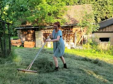 A woman with red hair in a light blue dress stands outside raking grass in front of a rustic barn on a sunny summer day.