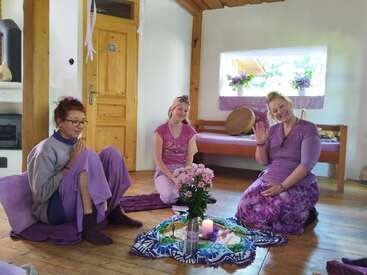 Three women dressed in purple sit together on the floor of a cozy, wooden room, gathered around flowers and candles, smiling and engaging in a peaceful activity.