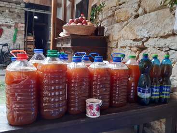 Large plastic bottles filled with homemade juice or cider line a rustic wooden table. A floral cup and bottles of water stand nearby; apples in a basket behind.
