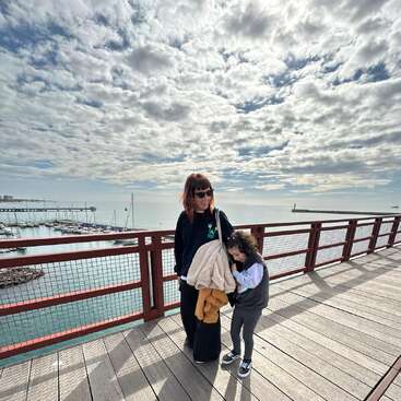 A woman and a child stand on a wooden pier above calm water, surrounded by boats, under a dramatic, cloud-filled sky. Both appear happy and relaxed.