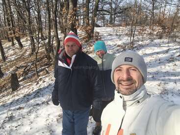 Three people dressed warmly are walking in a snowy forest. The sun is shining, and they appear to be enjoying a peaceful winter day outdoors together.