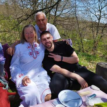 Three people sit outdoors enjoying a picnic. The woman wears a white embroidered dress, while the men smile and pose. Trees and nature surround them in spring.