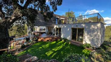 La imagen muestra el exterior de una casa moderna, con un gran jardín delantero con patio, entrada de vehículos y garaje, y un espacio exterior bien cuidado y acogedor.