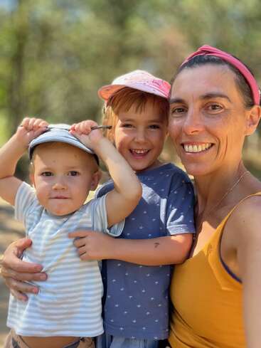 La imagen muestra a una mujer con top amarillo y diadema rosa posando con dos niños, uno con camisa azul y el otro con camisa de rayas, en un entorno natural al aire libre.