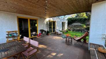 Un acogedor patio cubierto con muebles de madera, ventanas con marcos amarillos, plantas y sillas de jardín da a un exuberante césped rodeado de rocas y vegetación, creando un ambiente relajante.
