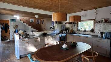 Esta imagen muestra una cocina acogedora y moderna con detalles en madera, lámparas colgantes, una mesa de comedor, una ventana grande, una escalera y varios electrodomésticos y objetos decorativos.