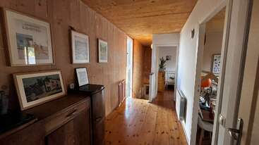 Un cálido pasillo de madera con cuadros en las paredes, suelos pulidos, muebles y puertas que dan a habitaciones luminosas, una de ellas con una planta y una cuna.