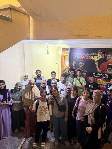 A group of smiling young people poses together indoors under a staircase. There’s a "Speak Up" language center sign behind them, and they look happy and friendly.