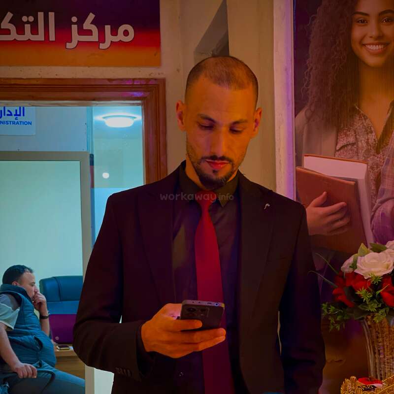 The image depicts a man in a suit holding a phone, standing in front of a poster and a doorway, with another man sitting in the background.