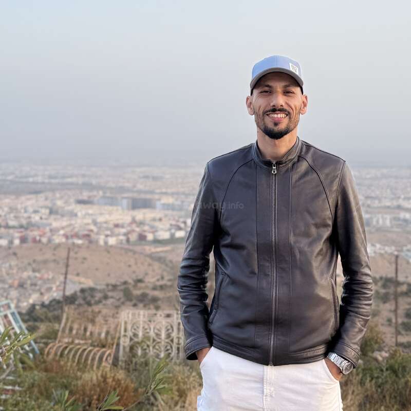 A man wearing a black leather jacket, white pants, and a light blue cap stands outdoors, smiling, with a cityscape and hills visible in the background.