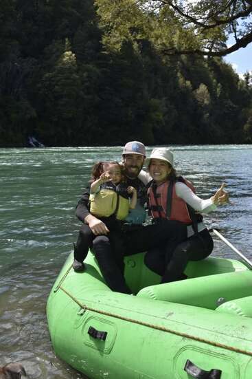 A joyful family sits on a green inflatable raft by a river, smiling and enjoying nature. They're wearing life jackets, surrounded by lush green trees.