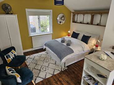 This cozy bedroom features a double bed with blue and white bedding, accent chairs, a geometric rug, yellow accent wall, shelves, and plenty of natural light.