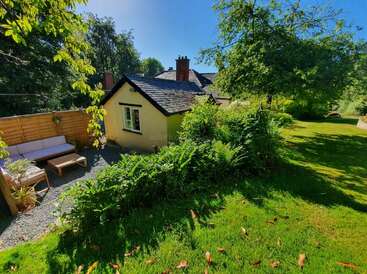 A charming cottage sits nestled among lush greenery under a clear blue sky. Outdoor seating, flowering plants, and bright sunlight create a peaceful, inviting garden scene.