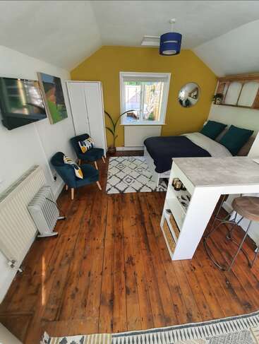 This cozy room features a yellow accent wall, wooden floors, a double bed, two chairs, a TV, wardrobe, rugs, plants, and modern decor.