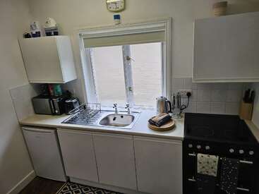 This compact kitchen features a sink, stove, and fridge. There’s a microwave, dish rack, kettle, bread, and utensils neatly arranged on clean white countertops.