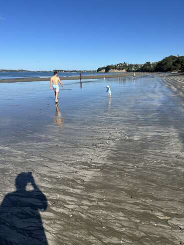 Un día soleado en la playa, con gente paseando y jugando cerca de la orilla. Aguas tranquilas, cielo azul, casas en la lejanía y la sombra del fotógrafo en primer plano.