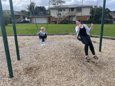 Una mujer y un niño pequeño se divierten columpiándose juntos en un parque infantil, rodeados de virutas de madera, césped verde y casas residenciales al fondo.
