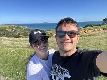 Una pareja sonriente se hace un selfi al aire libre en un día soleado, con gafas de sol, con colinas verdes, el mar azul y un cielo despejado como pintoresco telón de fondo.