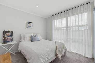 Se trata de un dormitorio luminoso y minimalista, con una cama blanca con cojines, una mesita de noche, cortinas transparentes en los grandes ventanales, paredes en un suave tono gris y suelo de moqueta.