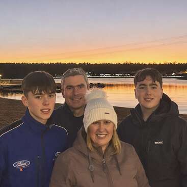 Uma família de quatro pessoas sorri à beira de um lago ao pôr do sol. Estão vestindo casacos de inverno e chapéus, bem juntinhos, com um lindo céu alaranjado ao fundo.