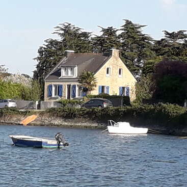 A charming stone house with blue shutters sits by the water, surrounded by trees. Small boats float gracefully on the calm river near the shore.