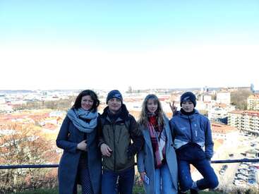 La imagen muestra a cuatro personas posando para una foto en una colina desde la que se domina una ciudad, con un cielo azul y edificios de fondo.
